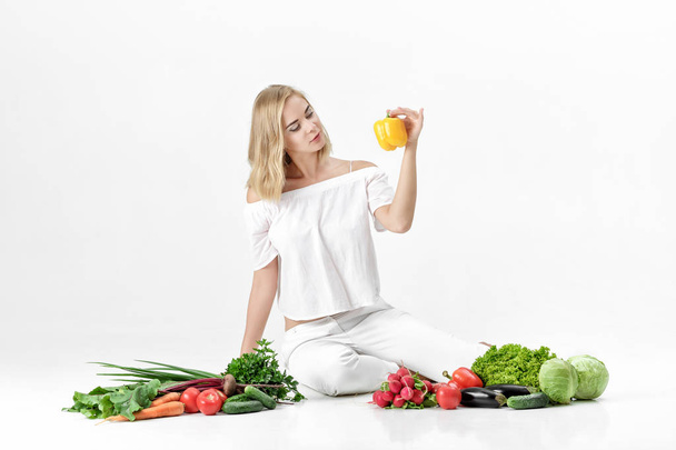Beautiful blond woman in white clothes and lots of fresh vegetables on white background. Girl holds bell pepper - Fotoğraf, Görsel