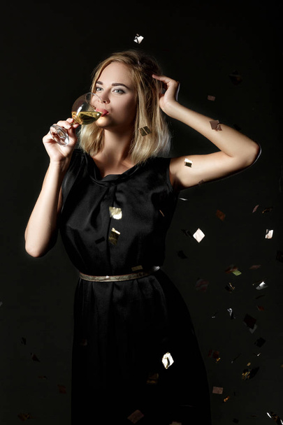 Beautiful happy blonde woman holding glass of white wine on black background with confetti - Photo, image