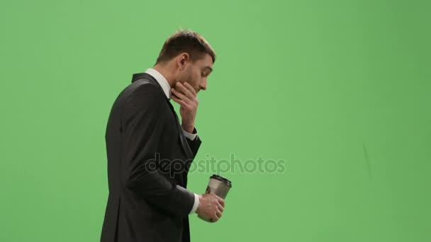 Tired businessman in a suit is walking with a coffee on a mock-up green screen in the background. - Video