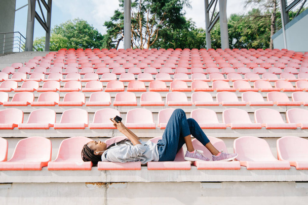 Horizontal view of the afro-american teenager laying on the seats on the stadium and chatting via the mobile phone. - Фото, зображення
