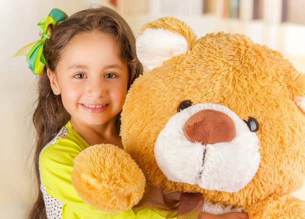 A portrait of a young pretty girl smiling and hugging her teddy bear over blurred background - Fotografie, Obrázek