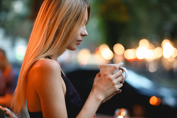 Woman sitting in a cafe drinking delicious latte. Coffee break outdoors in evening. - Photo, Image