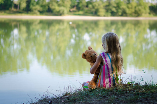 criança com ursinho sentado junto ao lago
 - Foto, Imagem