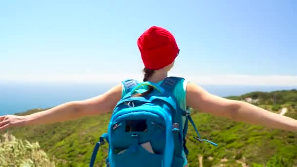 Una mujer con una mochila detrás de su espalda gira sus brazos hacia los lados, de pie en la cima de una colina en la orilla del océano
 - Imágenes, Vídeo