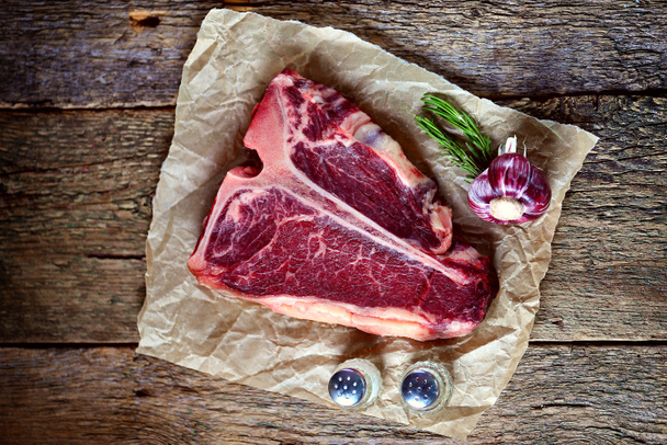 rohes T-Bone-Steak mit Knoblauch und Rosmarin auf altem Holzboden. rustikaler Stil. - Foto, Bild