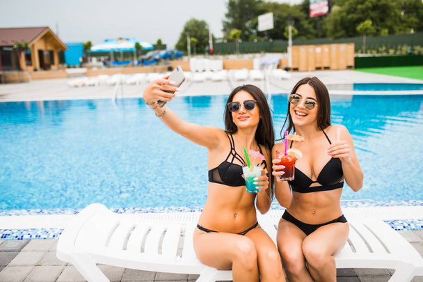Two attractive brunette women wearing bikini posing near the swimming pool, making selfie photo. Summer time - Zdjęcie, obraz