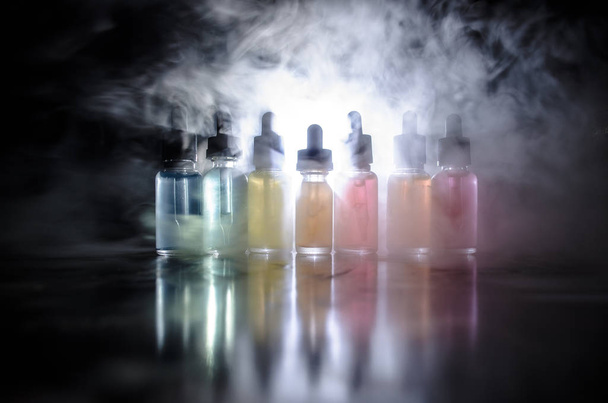 Η ιδέα της ταινίας. Σύννεφα καπνού και υγρά μπουκάλια σε σκούρο φόντο. Εφέ φωτός. Χρήσιμο ως background ή vape διαφήμιση ή vape φόντο. Επιλεκτική εστίαση - Φωτογραφία, εικόνα