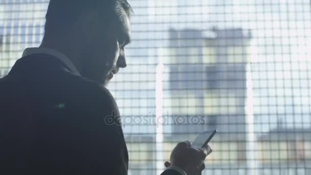Businessman in a suit is using a smartphone next to a big window in a garage. - Кадри, відео