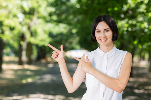 Kobieta uśmiechający się wskazujący obiema rękami. Pewnie kobieta na tle zielonego parku. Sukces i Ładna Brunetka Kobieta.  - Zdjęcie, obraz