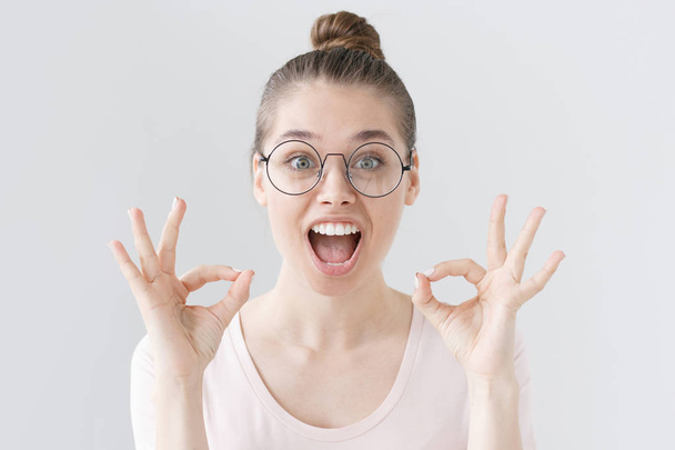 Photo of young European woman isolated on grey background in thin-framed eyeglasses with emotional expression of surprise and joy, her mouth and eyes wide open as she sees unbelievable things. - Fotoğraf, Görsel