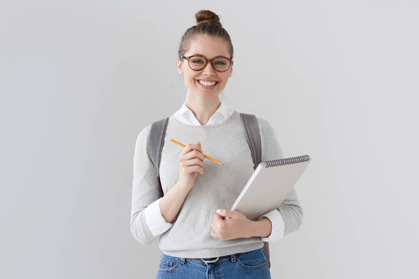 Indoor portrait of nice young European woman in hipster glasses standing isolated on gray background with big notebook in hands and pencil ready to note new information for studies or freelancing. - Fotó, kép