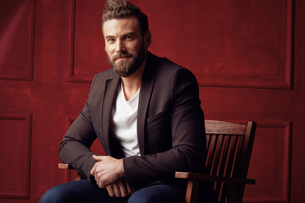 Bonito homem de barba sorridente, vestindo camisa branca e casaco marrom, sentado em cadeira de madeira em estúdio vermelho escuro
  - Foto, Imagem