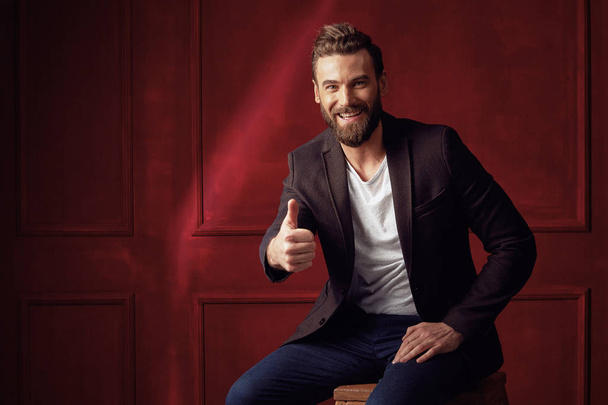 Attractive young beautiful and confident bearded man, wearing white shirt and brown jacket, sitting on wooden chair in studio with dark red background and showing his thumb up and smiling - Fotoğraf, Görsel
