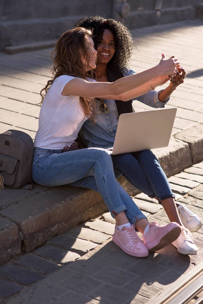 Two girls sitting on the pavement with the laptop and making selfie. One girl is black. Girls wearing casual clothes. - Fotoğraf, Görsel
