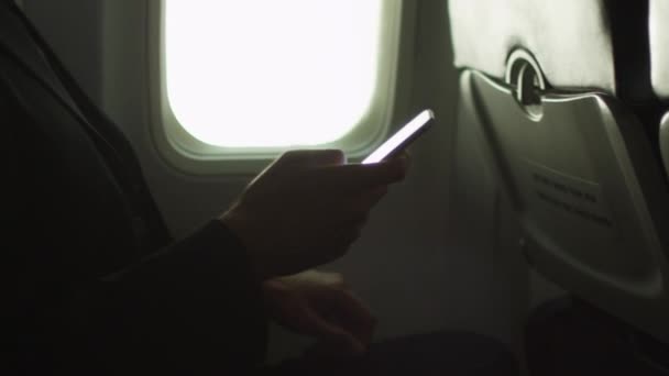 Young man in close-up is using a smartphone inside an airplane next to a window. - Πλάνα, βίντεο