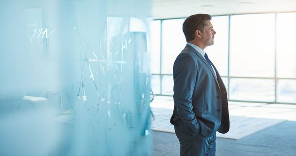 Ώριμες επιχειρηματία σε ένα εταιρικό κοστούμι στέκεται σε ένα χώρο μεγάλο κενό γραφείο με μοντέρνο γυάλινο διαχωριστικό δωματίου και αναζητούν μακριά από τα μεγάλα παράθυρα αισιοδοξία - Φωτογραφία, εικόνα
