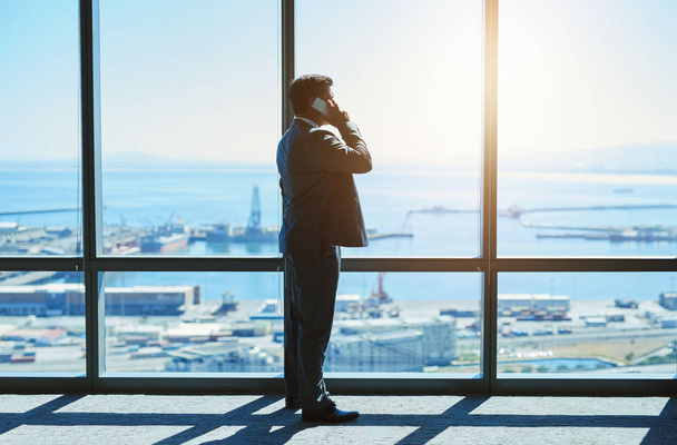 Volwassen bedrijfsleider staat naast grote ramen in een kantoor op de bovenste verdieping kijkend naar het uitzicht op de haven terwijl hij logistiek praat op zijn mobiele telefoon. - Foto, afbeelding