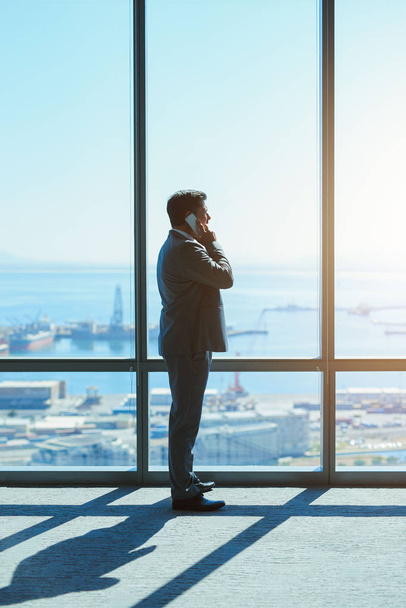 Mature business leader standing and looking out of large windows in a top floor office, talking on his mobile phone while looking out at the view - Foto, Imagem