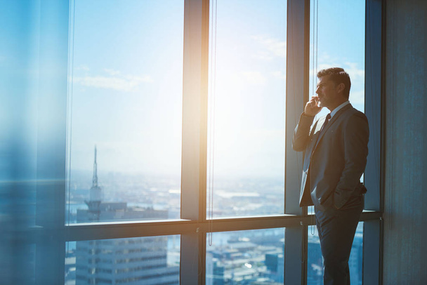 Handsome mature businessman standing in a professional corporate suit alongside large windows with a view of the city, talking on his mobile phone and looking out  - Foto, imagen