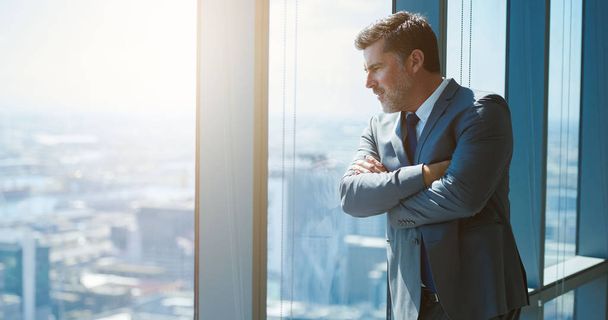 Handsome mature business executive with attractive designer stubble, standing with his arms crossed and leaning against a large window in a top floor office, looking down at city - Photo, Image