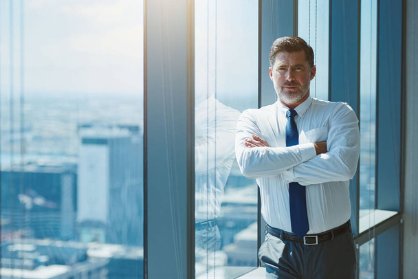 Portrait of a mature and handsome business executive looking seriously at the camera with his arms crossed, while leaning against a large window with a city view beyond - Fotó, kép