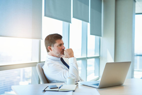 Knap volwassen business manager zitten aan zijn bureau in een modern kantoor met zijn laptop open voor hem weg te kijken met een zeer doordachte uitdrukking. - Foto, afbeelding
