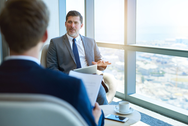 Handvoll reifer Unternehmensmanager sitzt in einem modernen Büroraum und hält ein digitales Tablet in der Hand, während er einen jungen Bewerber interviewt - Foto, Bild