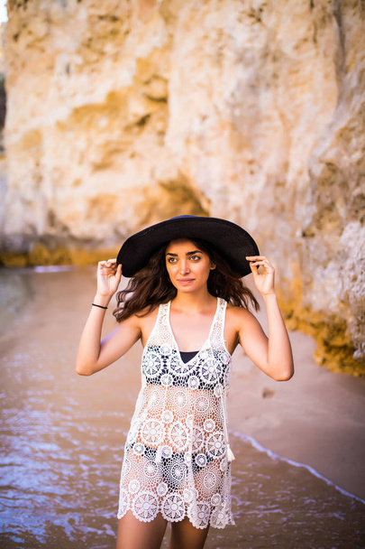 Portrait of beautiful latin girl on the beach with black hat smile neat ocean and rocks  on summer vocation - Foto, Imagem