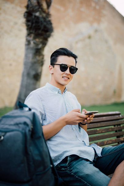 Молодий азіатський чоловік в окулярах сидить на лавці і дивиться за його телефоном
 - Фото, зображення