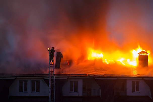 Brennende Stichflamme mit Rauch auf dem Dach eines Mehrfamilienhauses in der Stadt, Feuerwehrmann oder Feuerwehrmann auf der Leiter löscht Brand. - Foto, Bild