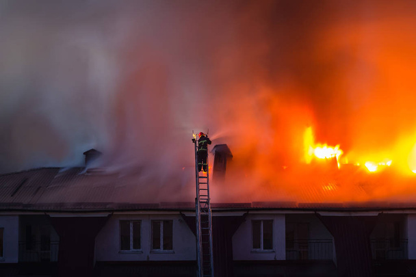 Płonący ogień z dymem na dachu kamienicy w mieście, strażak lub strażak na drabinie gaśnie pożar. - Zdjęcie, obraz