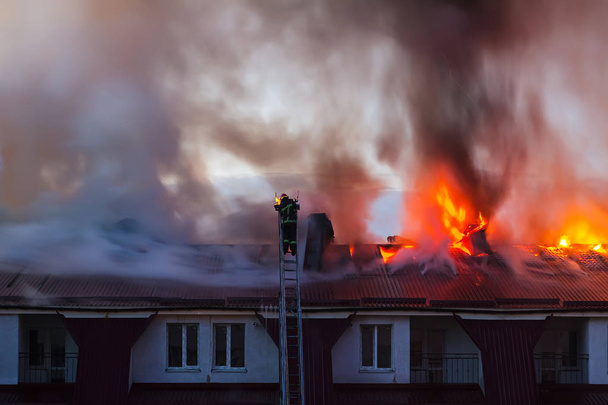 Burning fire flame with smoke on the apartment house roof in the city, firefighter or fireman on the ladder extinguishes fire. - Photo, Image