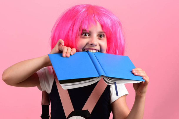 Pupil with pink wig. Girl bites big blue book. - Foto, imagen