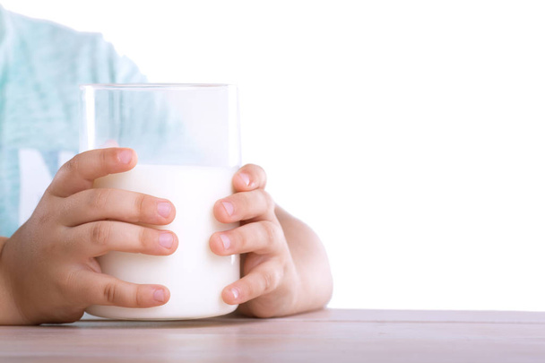 The small fingers of a little kid are holding a large glass of milk, which stands on a wooden table isolated on a white background. - Foto, immagini