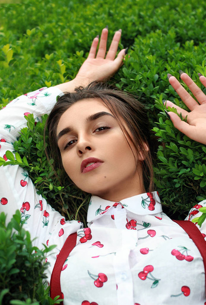 Closeup stylish beautiful hipster girl laying in boxwood bush. Young girl with beautiful face in spring. space for text. Joyful moment.Brunette girl in shirt with print chery and braces for trousers - Foto, imagen