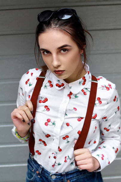 Closeup fashion portrait of young pretty hipster girl with dark brown hairs wearing stylish ,denim braces for trousers glasses and shirt with funny print cherry .Doors of garage bakground - Foto, Imagen