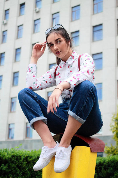 Fashion portrait of young pretty hipster girl with dark brown hairs wearing stylishnim braces for trousers glasses and shirt with funny print cherry siting on yellow dump.Urban style background - Photo, Image