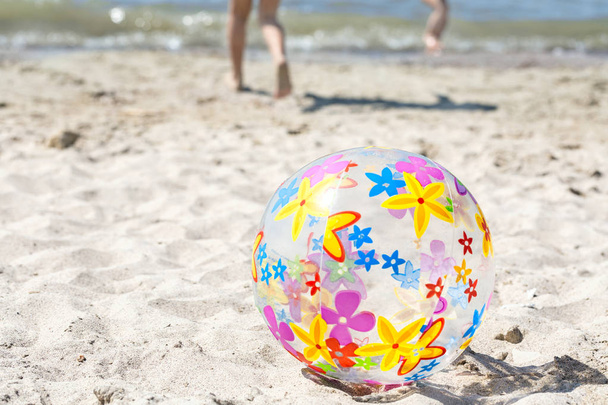 Ένα πολύχρωμο φουσκωτή μπάλα βρίσκεται πάνω στην παραλία στην άμμο. Το παιδί τρέχει δίπλα από τη φουσκωτή μπάλα. Καλοκαιρινή διασκέδαση έννοια. - Φωτογραφία, εικόνα
