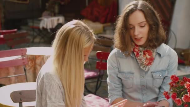 Dwie dziewczyny są rozmowy i za pomocą tabletu w odkrytym Coffee Shop - Materiał filmowy, wideo
