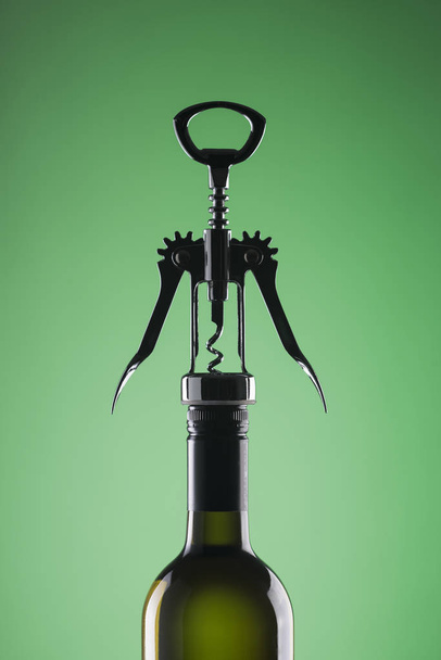 Een fles wijn met een stijlvolle kurkentrekker voor opening op een groene achtergrond. - Foto, afbeelding