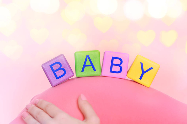 Color cubes with word BABY on abdomen of pregnant woman against Christmas lights background - Fotografie, Obrázek