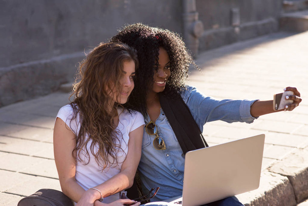Two happy girls sitting on the pavement with the laptop and making selfie. One girl is black. - Foto, Imagen