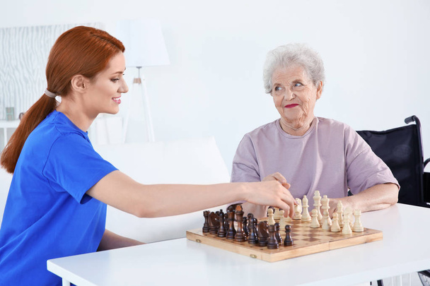 Young female volunteer playing chess with elderly woman in light room - Fotoğraf, Görsel