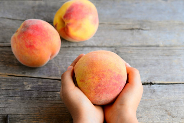 Small child holds a ripe peach in his hands. Sweet delicious peaches on an old wooden table. Encouraging healthy eating habits for kids - Photo, Image