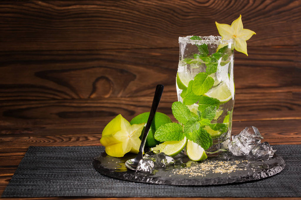 A huge glass filled with alcohol beverage from juicy lime, rum, fresh mint, decorative carambola and crushed ice on a dark wooden background. Summer mojito. Copy space. - Photo, Image