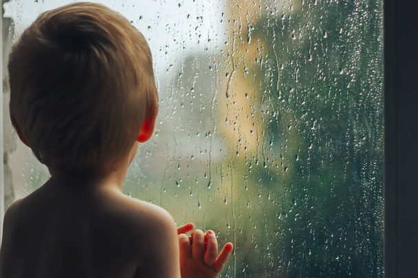μωρό ψάχνει στη βροχή μέσα από το παράθυρο που στέκεται με την πλάτη - Φωτογραφία, εικόνα