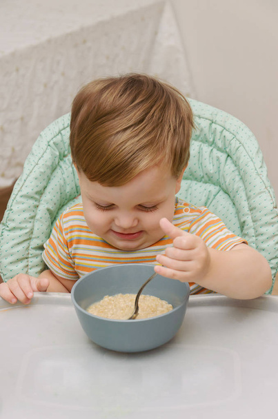 μικρό ξανθό αγόρι σε ένα ριγέ μπλουζάκι τρώει το χυλό με ένα κουτάλι - Φωτογραφία, εικόνα