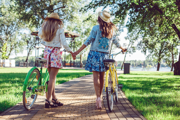 Rear view of two women holding hands walking in a park with bicycles. - 写真・画像