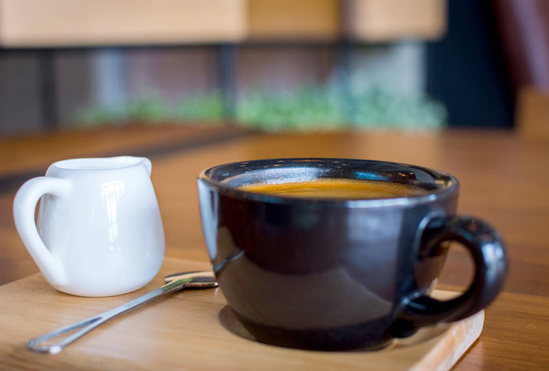 Dark black blue coffee cup with americano coffee, spoon and a milk jug on a wooden stand - Fotó, kép