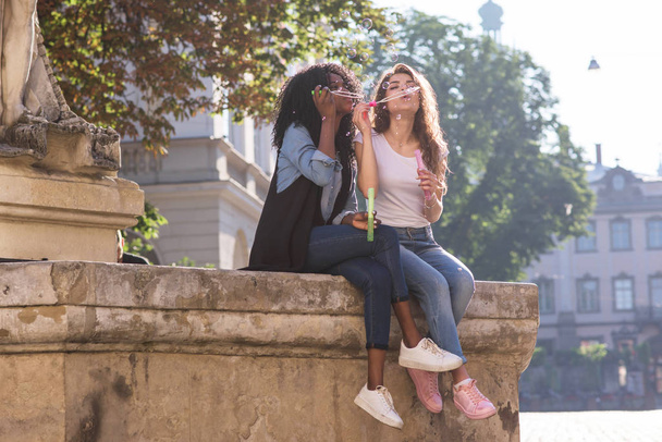 Bubble blowers sitting near the monument in the downtown. Girls wearing casual clothes. One girl is black. - Zdjęcie, obraz